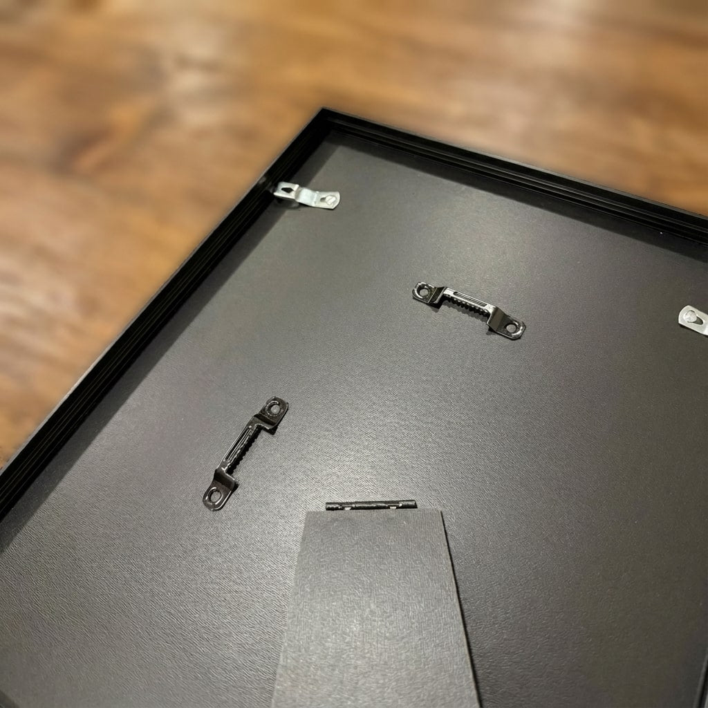 Close-up rear view of the high-quality black aluminum frame on a wooden table. Shows sturdy metal hanging hardware for wall mounting and a fold-out easel stand for desk display.