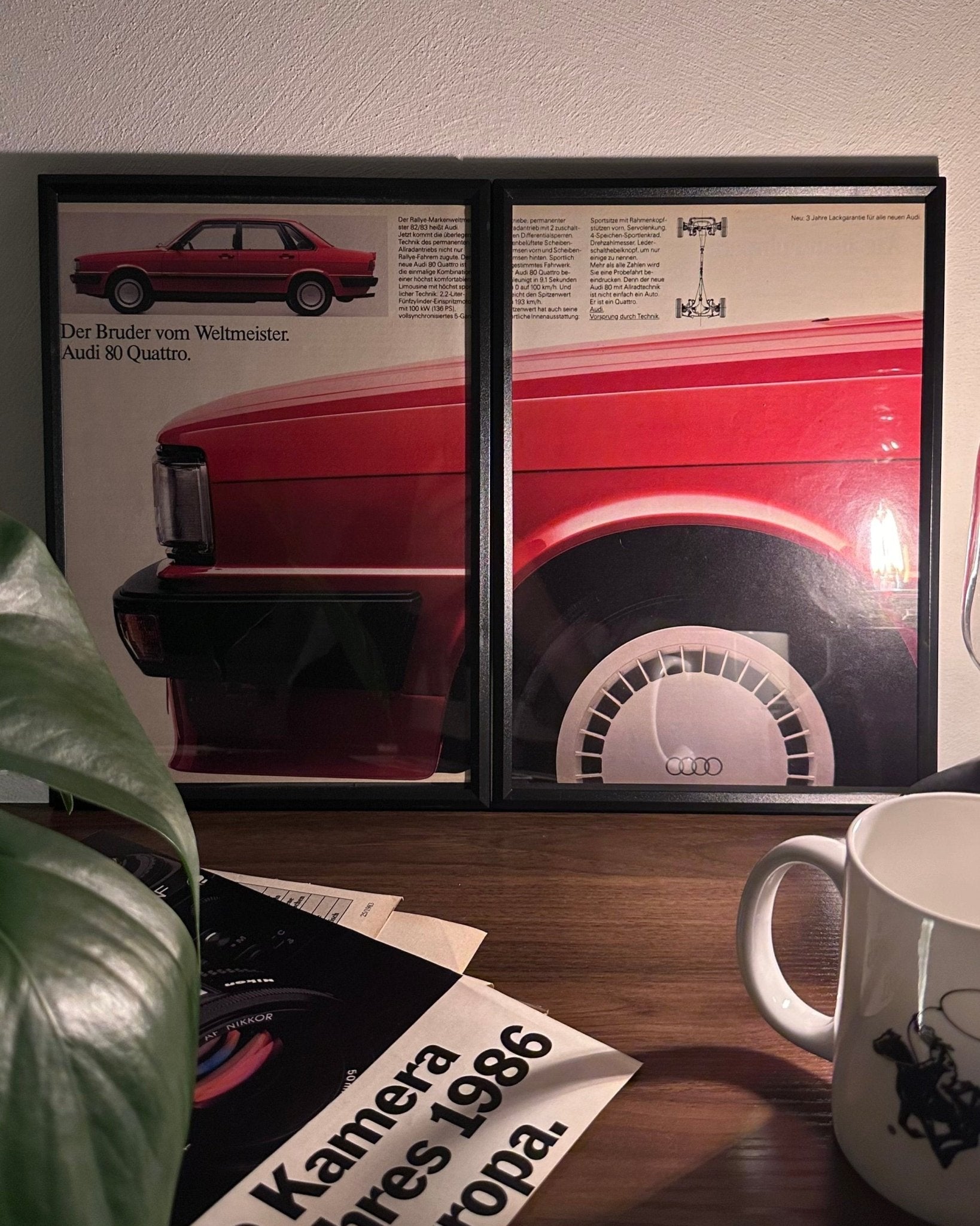 Gerahmte Oldtimer-Werbung des Audi 80 Quattro, stilvoll auf einem Tisch präsentiert, umgeben von Vintage-Magazinen und einer Tasse. Perfektes Ambiente, das die klassische Eleganz des Posters zur Geltung bringt.