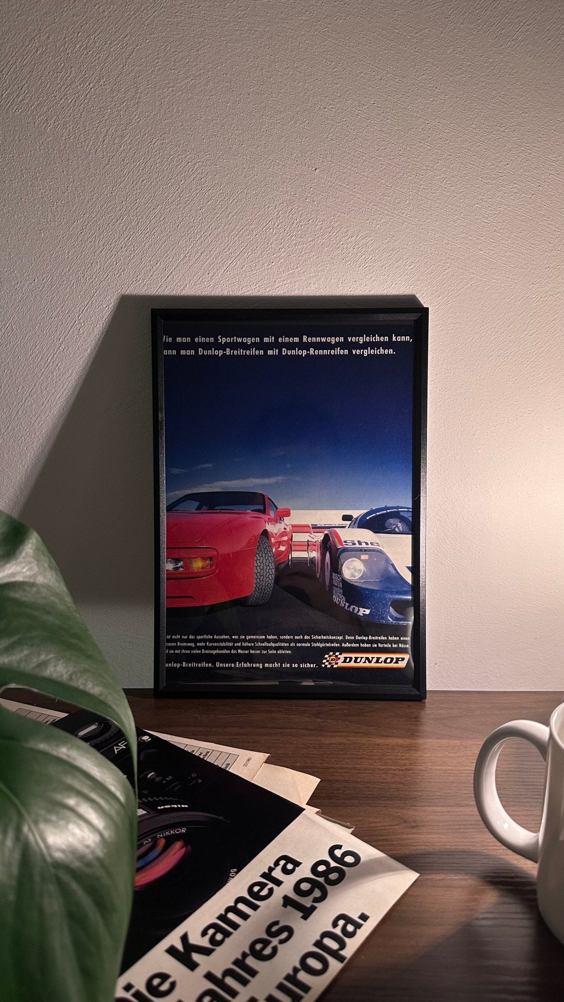 Gerahmte Oldtimer-Werbung des Porsche 956, stilvoll auf einem Tisch präsentiert, umgeben von Vintage-Magazinen und einer Tasse. Perfektes Ambiente, das die ikonische Le Mans Historie und die Rennsport-Dominanz des Porsche 956 zur Geltung bringt.