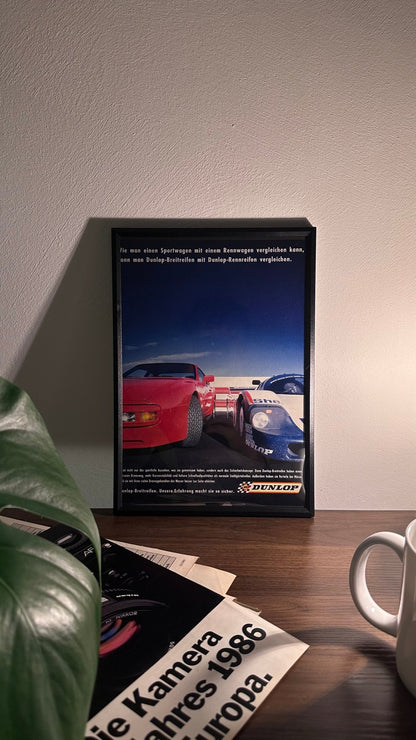 Gerahmte Oldtimer-Werbung des Porsche 956, stilvoll auf einem Tisch präsentiert, umgeben von Vintage-Magazinen und einer Tasse. Perfektes Ambiente, das die ikonische Le Mans Historie und die Rennsport-Dominanz des Porsche 956 zur Geltung bringt.