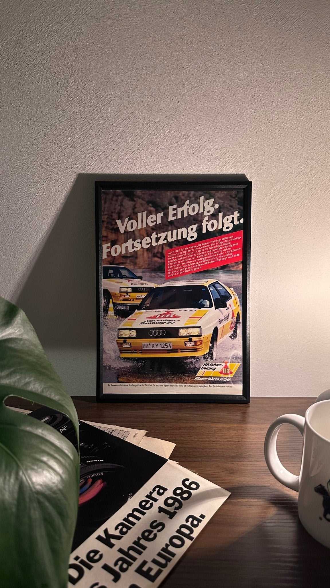 Gerahmte Oldtimer-Werbung des Audi Urquattro, stilvoll auf einem Tisch präsentiert, umgeben von Vintage-Magazinen und einer Tasse. Perfektes Ambiente, das die klassische Eleganz des Posters zur Geltung bringt.