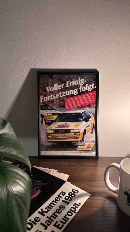 Gerahmte Oldtimer-Werbung des Audi Urquattro, stilvoll auf einem Tisch präsentiert, umgeben von Vintage-Magazinen und einer Tasse. Perfektes Ambiente, das die klassische Eleganz des Posters zur Geltung bringt.