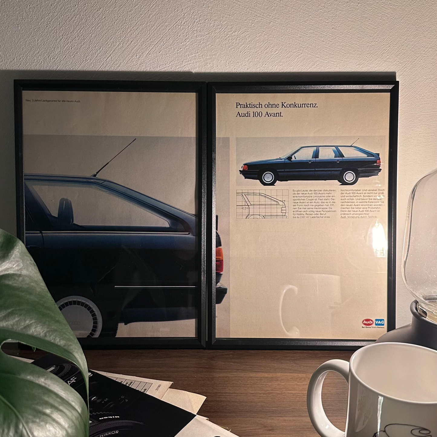 Gerahmte Oldtimer-Werbung des Audi 100 Avant, stilvoll auf einem Tisch präsentiert, umgeben von Vintage-Magazinen und einer Tasse. Perfektes Ambiente, das die klassische Eleganz des Posters zur Geltung bringt.