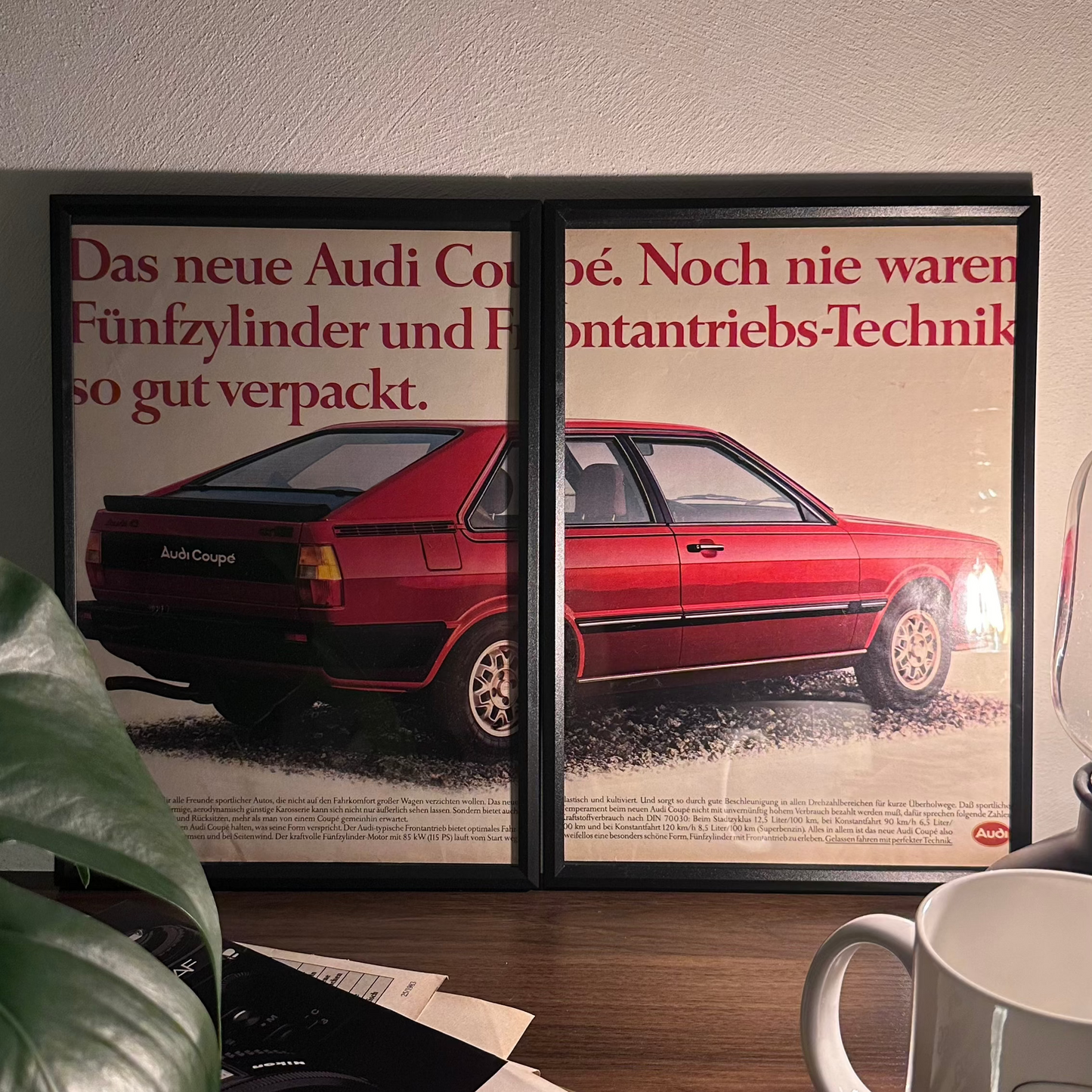 Gerahmte Oldtimer-Werbung des Audi 80 Coupé, stilvoll auf einem Tisch präsentiert, umgeben von Vintage-Magazinen und einer Tasse. Perfektes Ambiente, das die klassische Eleganz des Posters zur Geltung bringt."