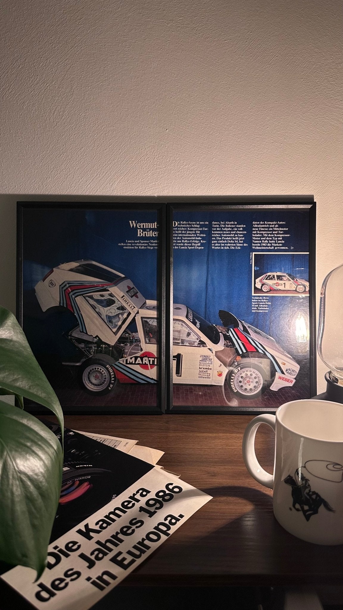 Gerahmte Oldtimer-Werbung des Lancia Delta S4, stilvoll auf einem Tisch präsentiert, umgeben von Vintage-Magazinen und einer Tasse. Perfektes Ambiente, das die klassische Eleganz des Posters zur Geltung bringt.