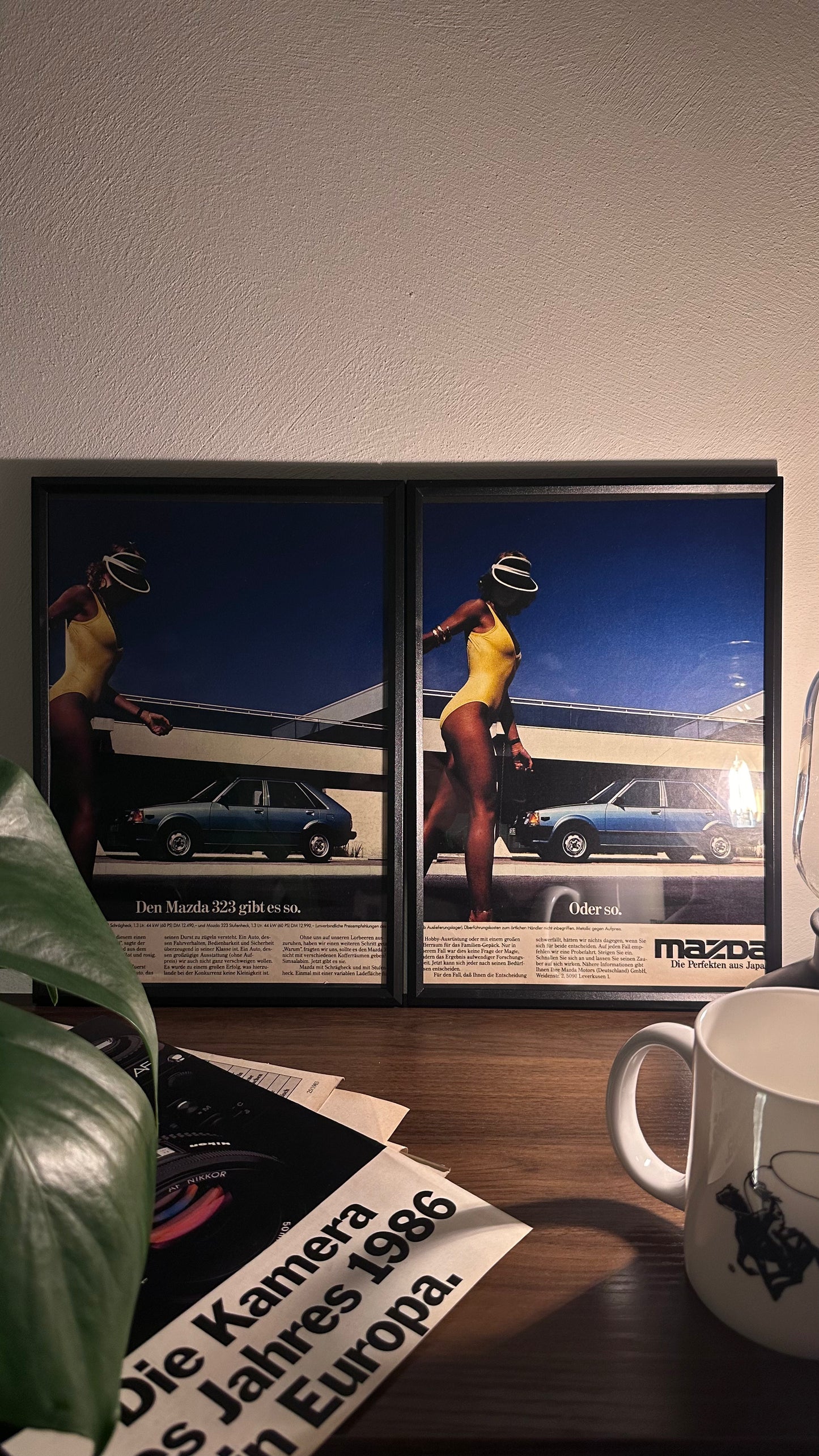 Gerahmte Oldtimer-Werbung des Mazda 323, stilvoll auf einem Tisch präsentiert, umgeben von Vintage-Magazinen und einer Tasse. Perfektes Ambiente, das die klassische Eleganz des Posters zur Geltung bringt.