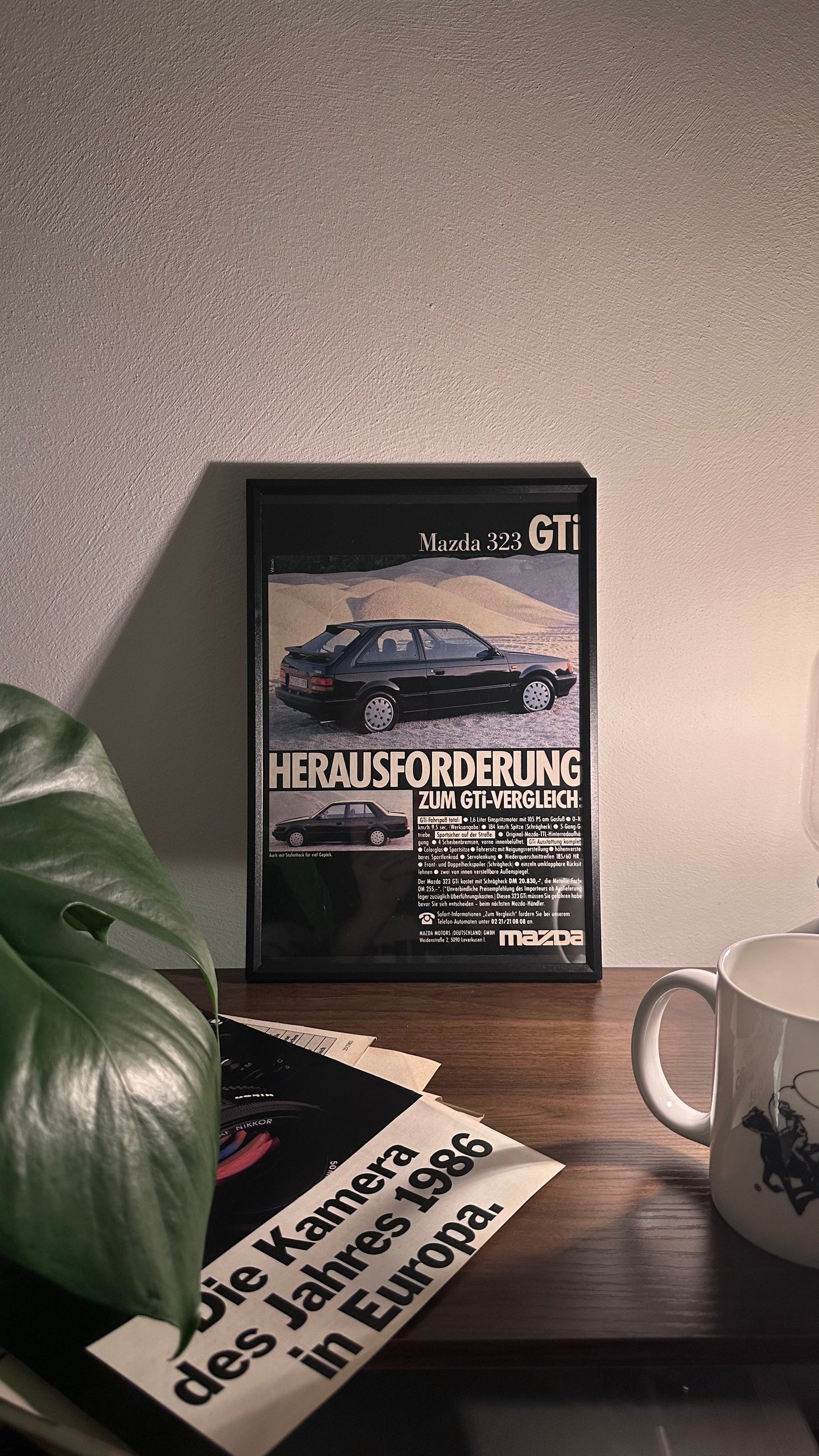 Gerahmte Oldtimer-Werbung des Mazda 323 GTI, stilvoll auf einem Tisch präsentiert, umgeben von Vintage-Magazinen und einer Tasse. Perfektes Ambiente, das die klassische Eleganz des Posters zur Geltung bringt.