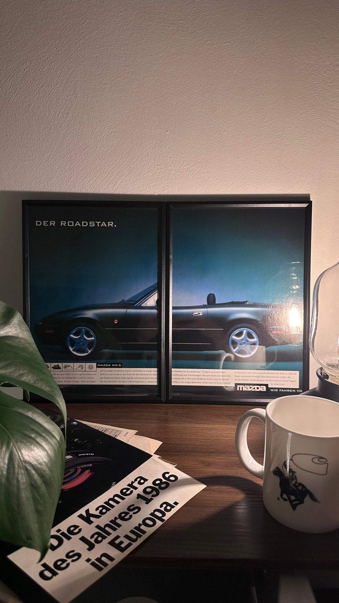 Gerahmte Oldtimer-Werbung des Mazda MX-5, stilvoll auf einem Tisch präsentiert, umgeben von Vintage-Magazinen und einer Tasse. Perfektes Ambiente, das die klassische Eleganz des Posters zur Geltung bringt.