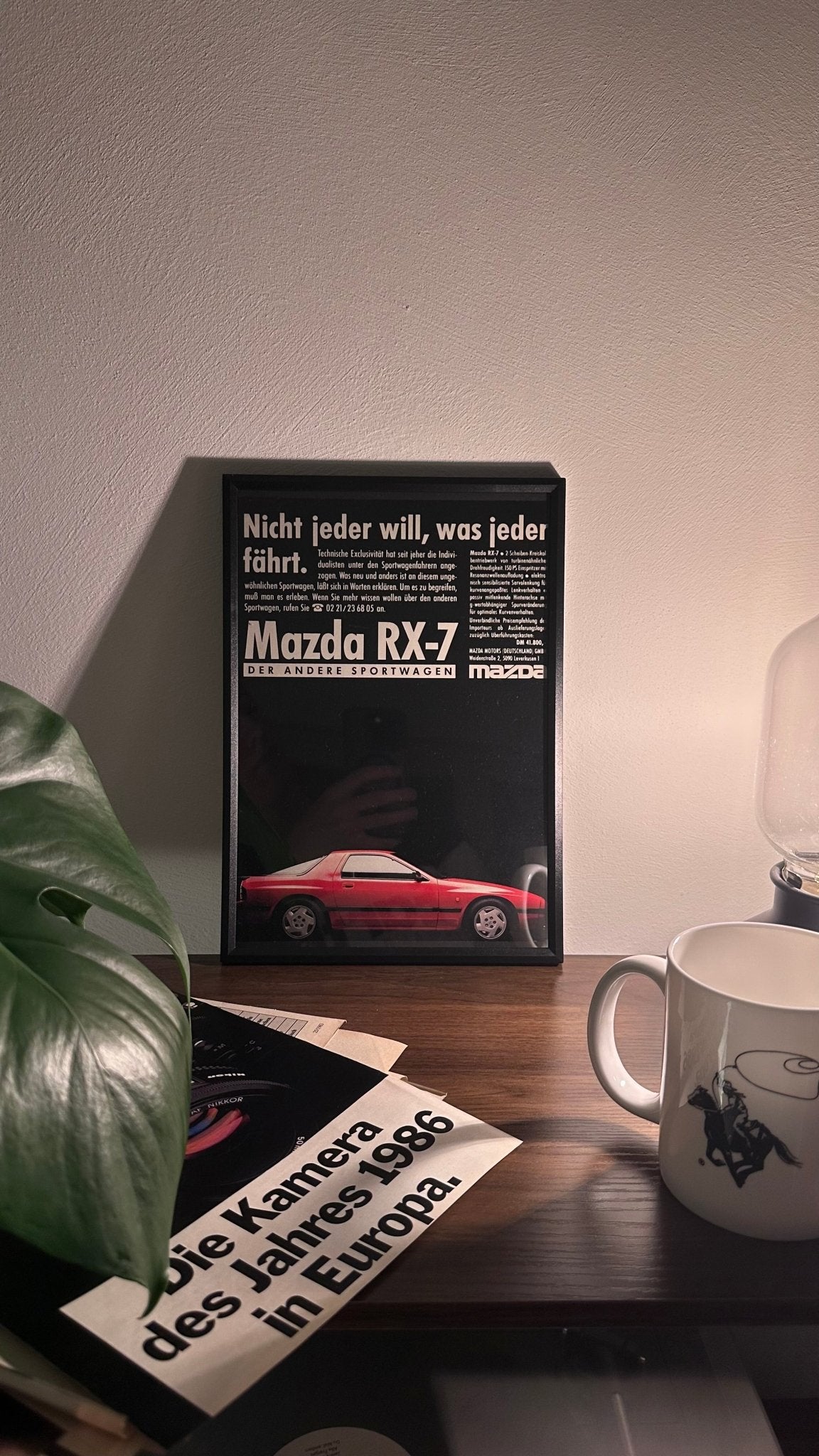 Gerahmte Oldtimer-Werbung des Mazda RX-7, stilvoll auf einem Tisch präsentiert, umgeben von Vintage-Magazinen und einer Tasse. Perfektes Ambiente, das die klassische Eleganz des Posters zur Geltung bringt.