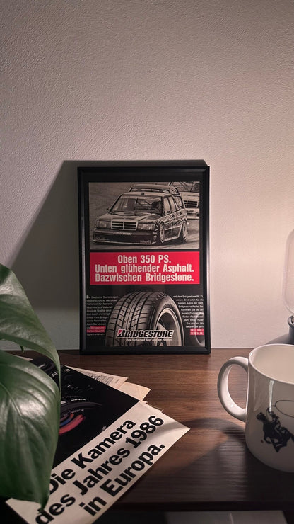 Gerahmte Oldtimer-Werbung des Mercedes-Benz 190 EVO2 DTM, stilvoll auf einem Tisch präsentiert, umgeben von Vintage-Magazinen und einer Tasse. Perfektes Ambiente, das die dynamische Eleganz des DTM-Klassikers zur Geltung bringt