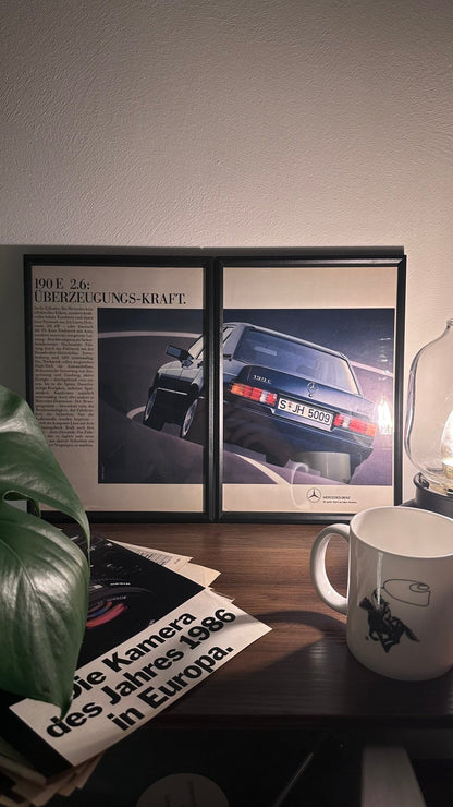 Gerahmte Oldtimer-Werbung des Mercedes-Benz 190E 2.6, stilvoll auf einem Tisch präsentiert, umgeben von Vintage-Magazinen und einer Tasse. Perfektes Ambiente, das die klassische Eleganz des Posters zur Geltung bringt.