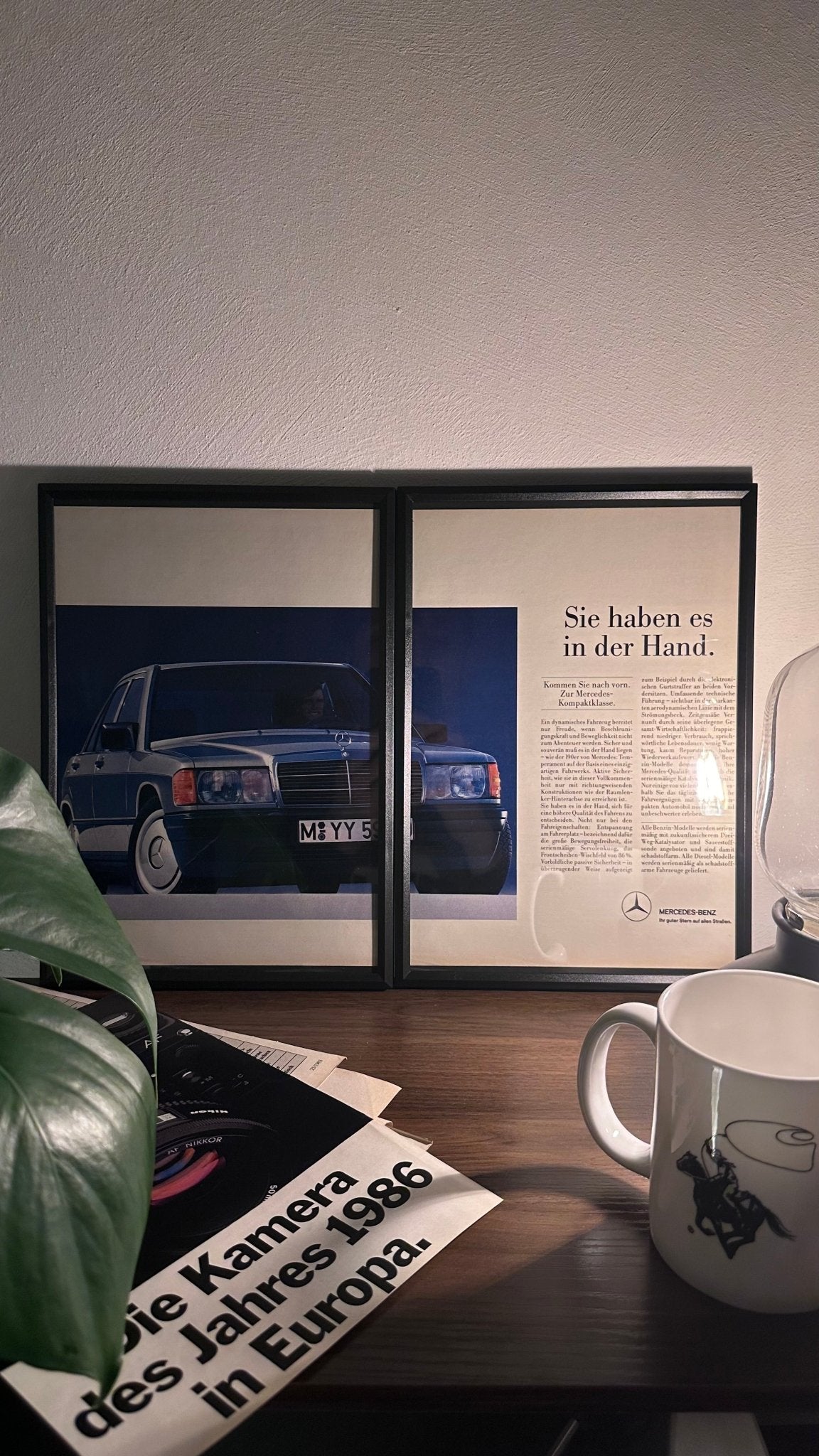 Gerahmte Oldtimer-Werbung des Mercedes-Benz 190E, stilvoll auf einem Tisch präsentiert, umgeben von Vintage-Magazinen und einer Tasse. Perfektes Ambiente, das die klassische Eleganz des Posters zur Geltung bringt