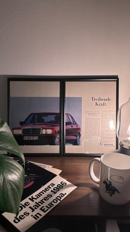 Gerahmte Oldtimer-Werbung des Mercedes-Benz 190E, stilvoll auf einem Tisch präsentiert, umgeben von Vintage-Magazinen und einer Tasse. Perfektes Ambiente, das die klassische Eleganz des Klassikers zur Geltung bringt.