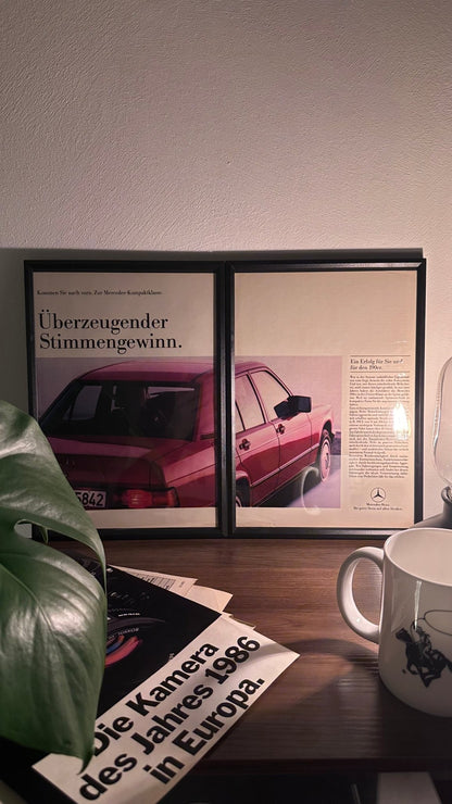 Gerahmte Oldtimer-Werbung des Mercedes-Benz 190E, stilvoll auf einem Tisch präsentiert, umgeben von Vintage-Magazinen und einer Tasse. Perfektes Ambiente, das die klassische Eleganz des Klassikers zur Geltung bringt.