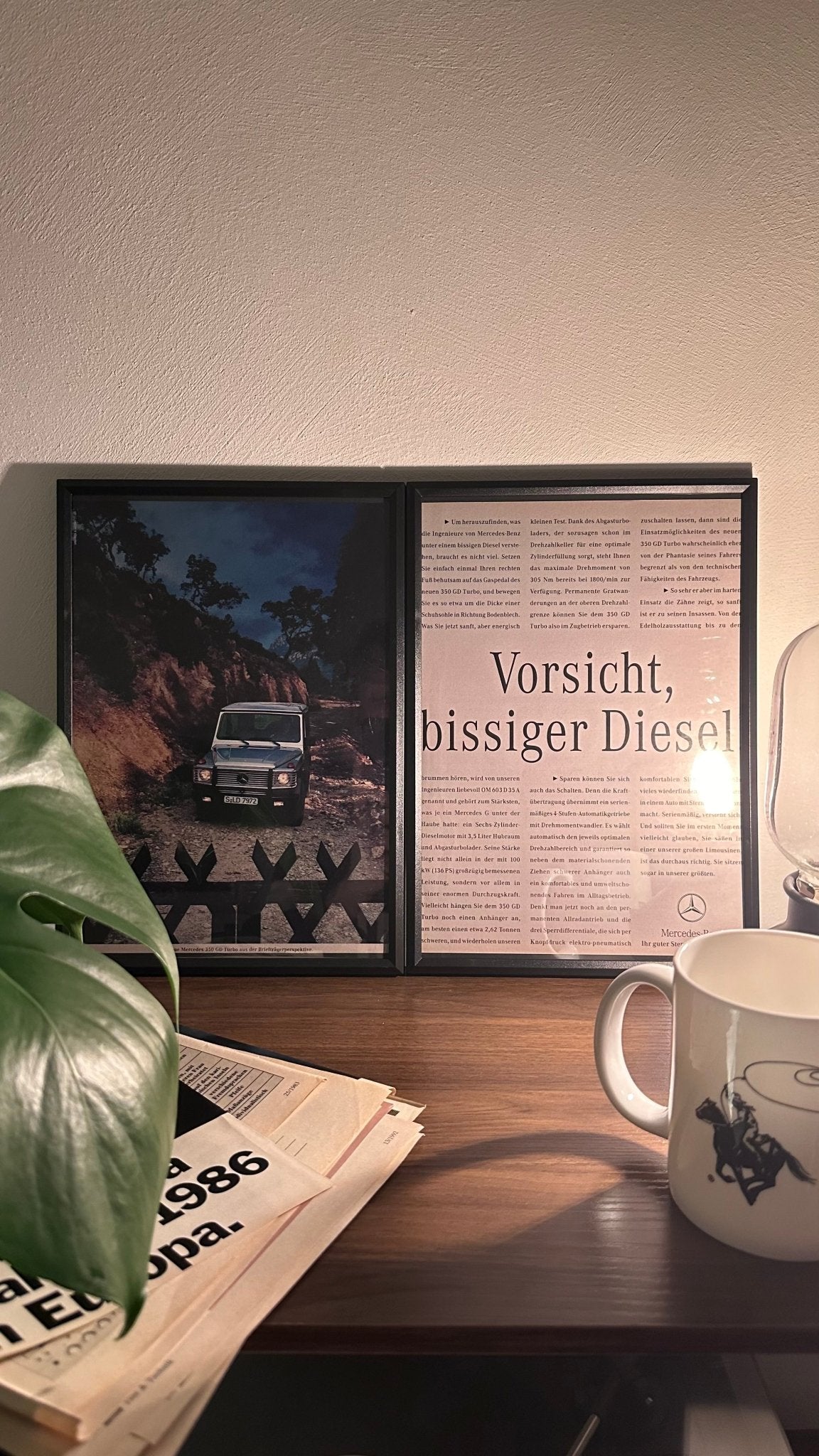 Gerahmte Oldtimer-Werbung des Mercedes-Benz 350 GD Turbo G-Klasse, stilvoll auf einem Tisch präsentiert, umgeben von Vintage-Magazinen und einer Tasse. Perfektes Ambiente, das die robuste Eleganz der G-Klasse zur Geltung bringt.