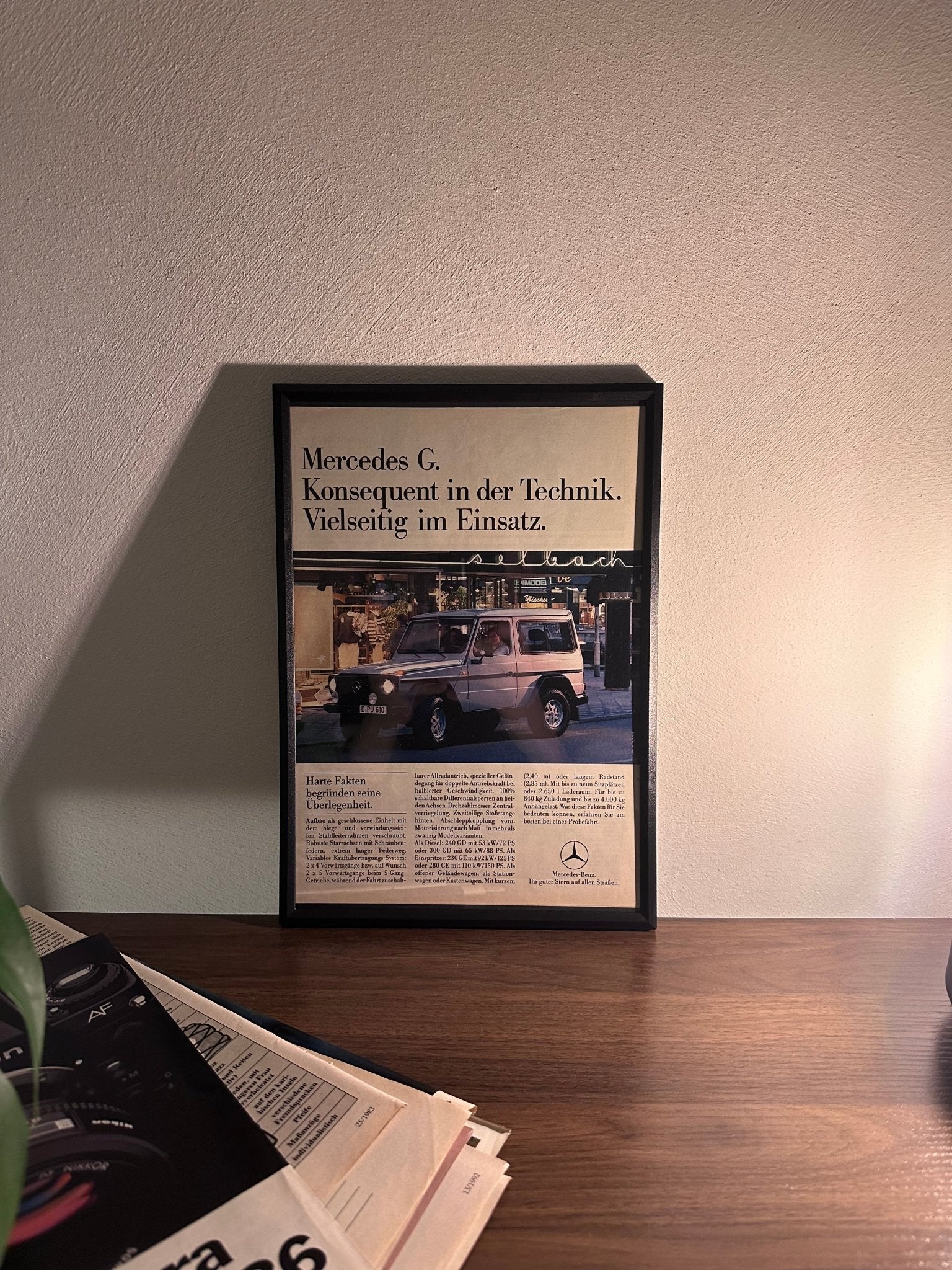 Gerahmte Oldtimer-Werbung der Mercedes-Benz G-Klasse, stilvoll auf einem Tisch präsentiert, umgeben von Vintage-Magazinen und einer Tasse. Perfektes Ambiente, das die robuste Eleganz der G-Klasse zur Geltung bringt.