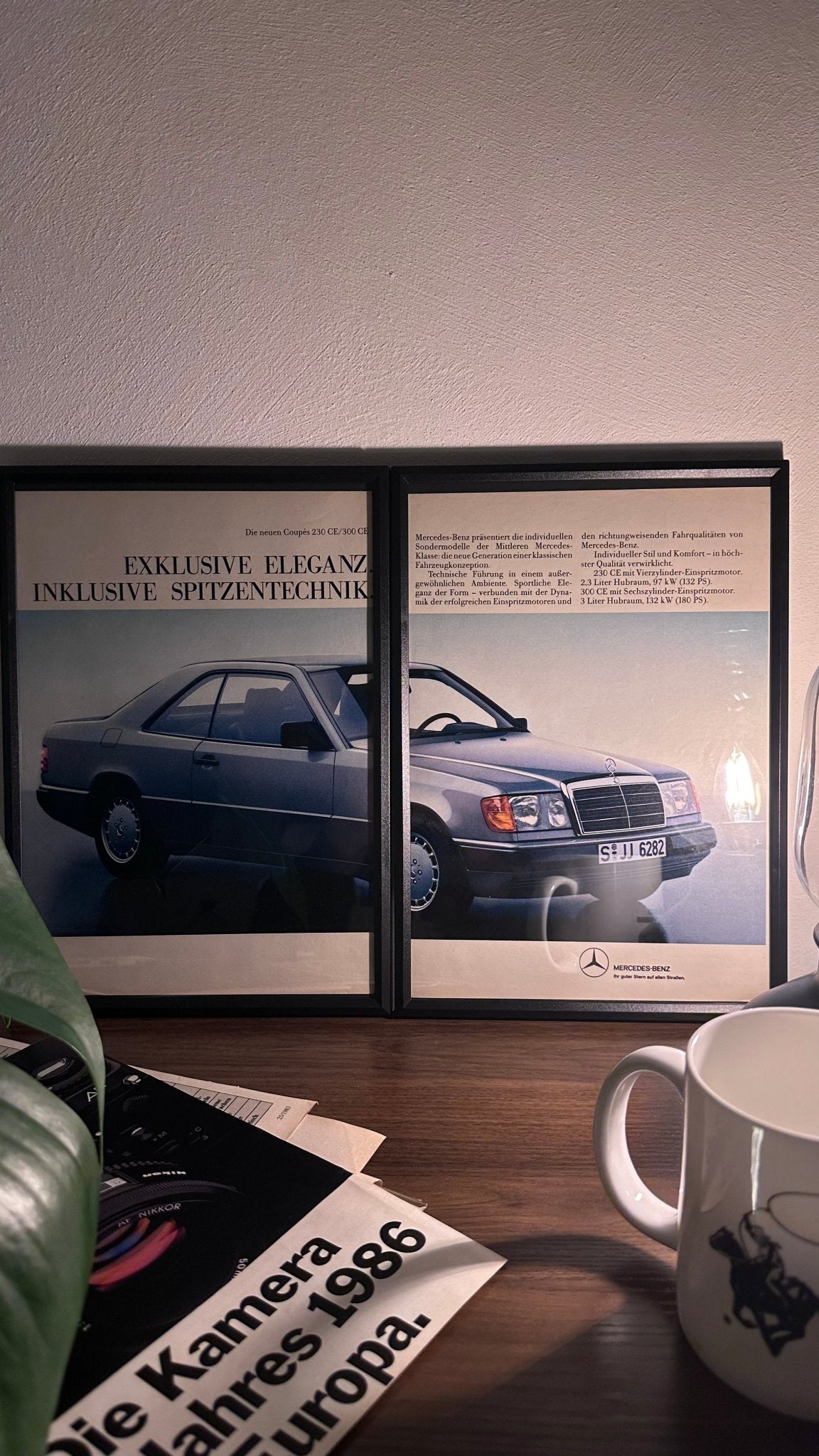 Gerahmte Oldtimer-Werbung des Mercedes-Benz W124 230/300 CE, stilvoll auf einem Tisch präsentiert, umgeben von Vintage-Magazinen und einer Tasse. Perfektes Ambiente, das die klassische Eleganz des Coupés zur Geltung bringt.