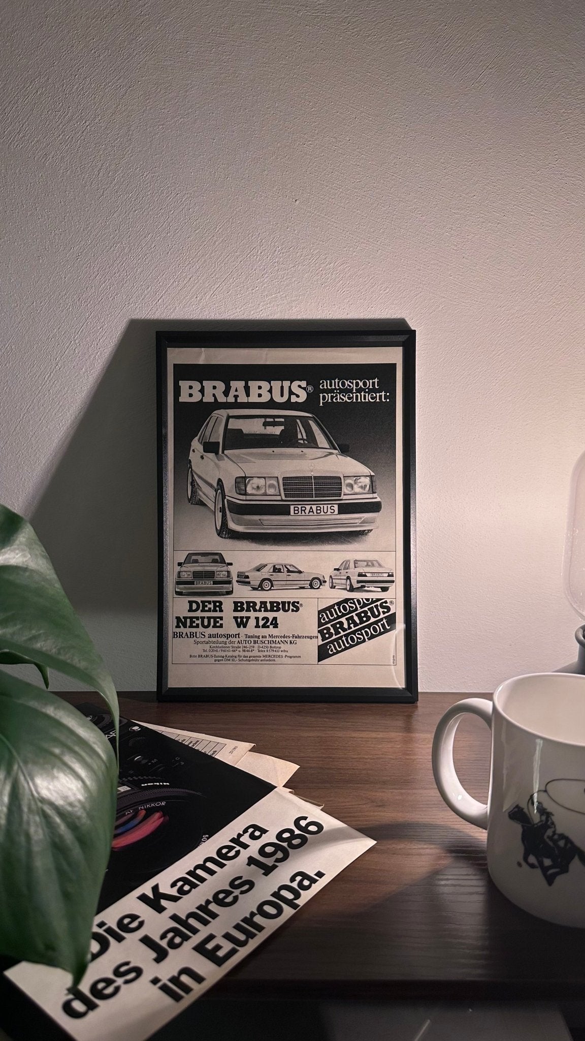 Gerahmte Oldtimer-Werbung des Mercedes-Benz W124 BRABUS, stilvoll auf einem Tisch präsentiert, umgeben von Vintage-Magazinen und einer Tasse. Perfektes Ambiente, das die kraftvolle Eleganz des BRABUS-getunten Klassikers zur Geltung bringt.