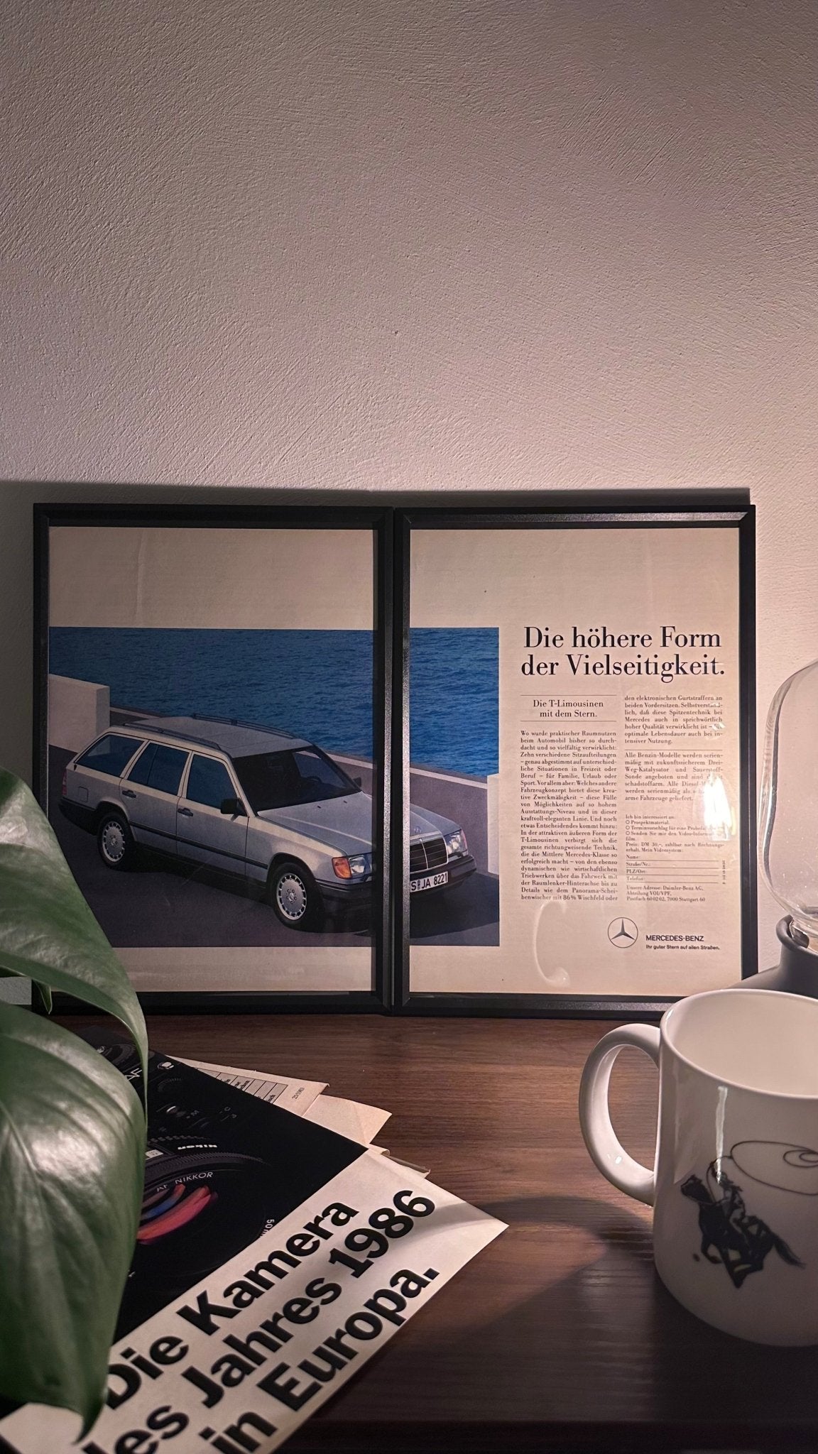 Gerahmte Oldtimer-Werbung des Mercedes-Benz W124 T-Modell, stilvoll auf einem Tisch präsentiert, umgeben von Vintage-Magazinen und einer Tasse. Perfektes Ambiente, das die klassische Eleganz des Kombi-Modells zur Geltung bringt.