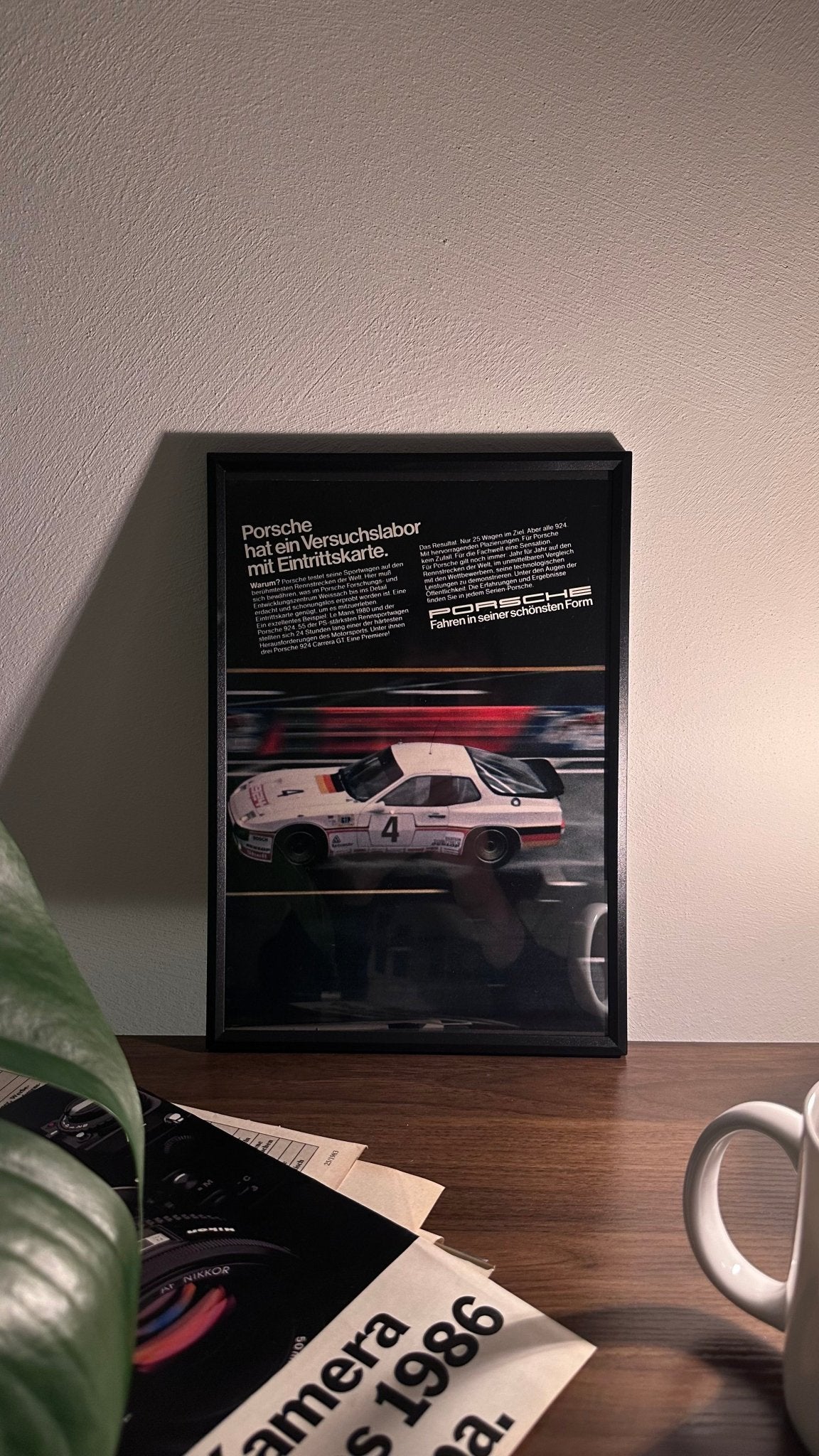 Gerahmte Oldtimer-Werbung des Porsche 924 Carrera GT, stilvoll auf einem Tisch präsentiert, umgeben von Vintage-Magazinen und einer Tasse. Perfektes Ambiente, das die sportliche Eleganz des Carrera GT zur Geltung bringt.