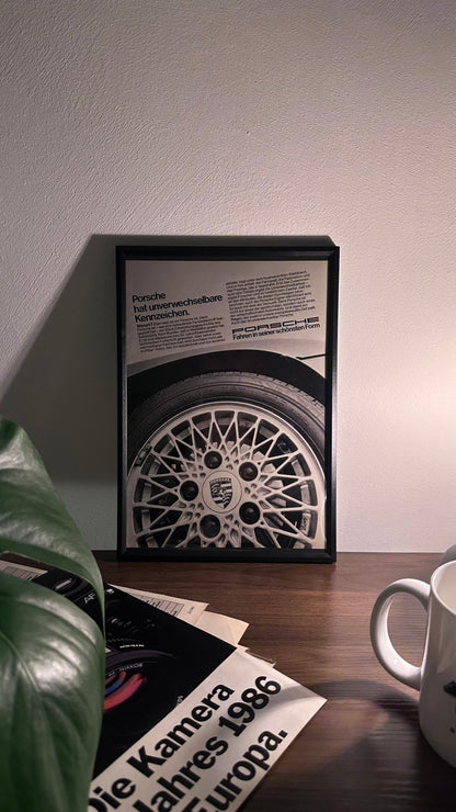 Gerahmte Oldtimer-Werbung der Porsche BBS Felge, stilvoll auf einem Tisch präsentiert, umgeben von Vintage-Magazinen und einer Tasse. Perfektes Ambiente, das die klassische Eleganz und Qualität der Porsche BBS Felgen zur Geltung bringt."