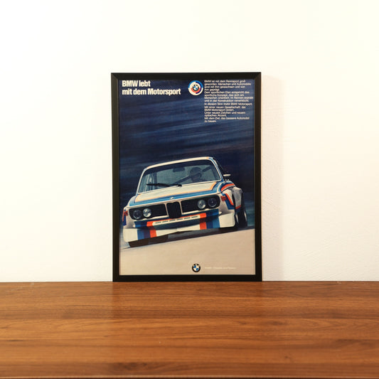 Original vintage BMW Motorsport advertisement framed in black aluminum. Single wall art piece showing the legendary BMW 3.0 CSL race car with M-stripes on a track, featuring the German headline 'BMW lebt mit dem Motorsport'.